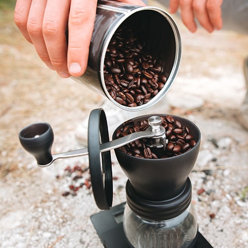 Adventure and Coffee — Person Filling Coffee Grinder with Fresh Beans at a Campsite Adventure and Coffee — Person Filling Coffee Grinder with Fresh Beans at a Campsite