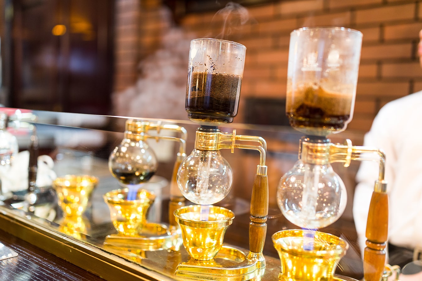 How to Use a Siphon Coffee Maker — A Siphon Coffee Maker With Gold and Wooden Accents Brewing Coffee in a Café