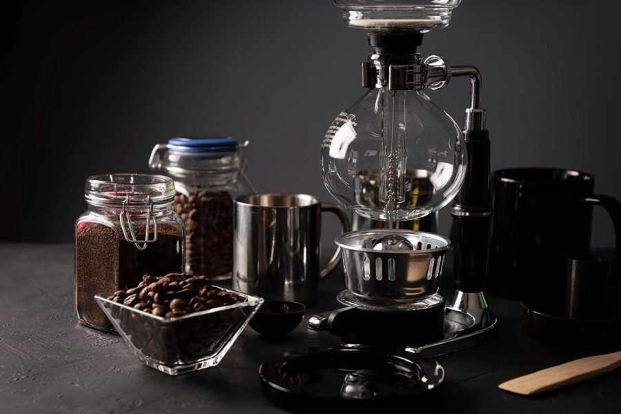 A Siphon Coffee Maker With Coffee Beans, Grounds, and Brewing Tools on a Dark Countertop
